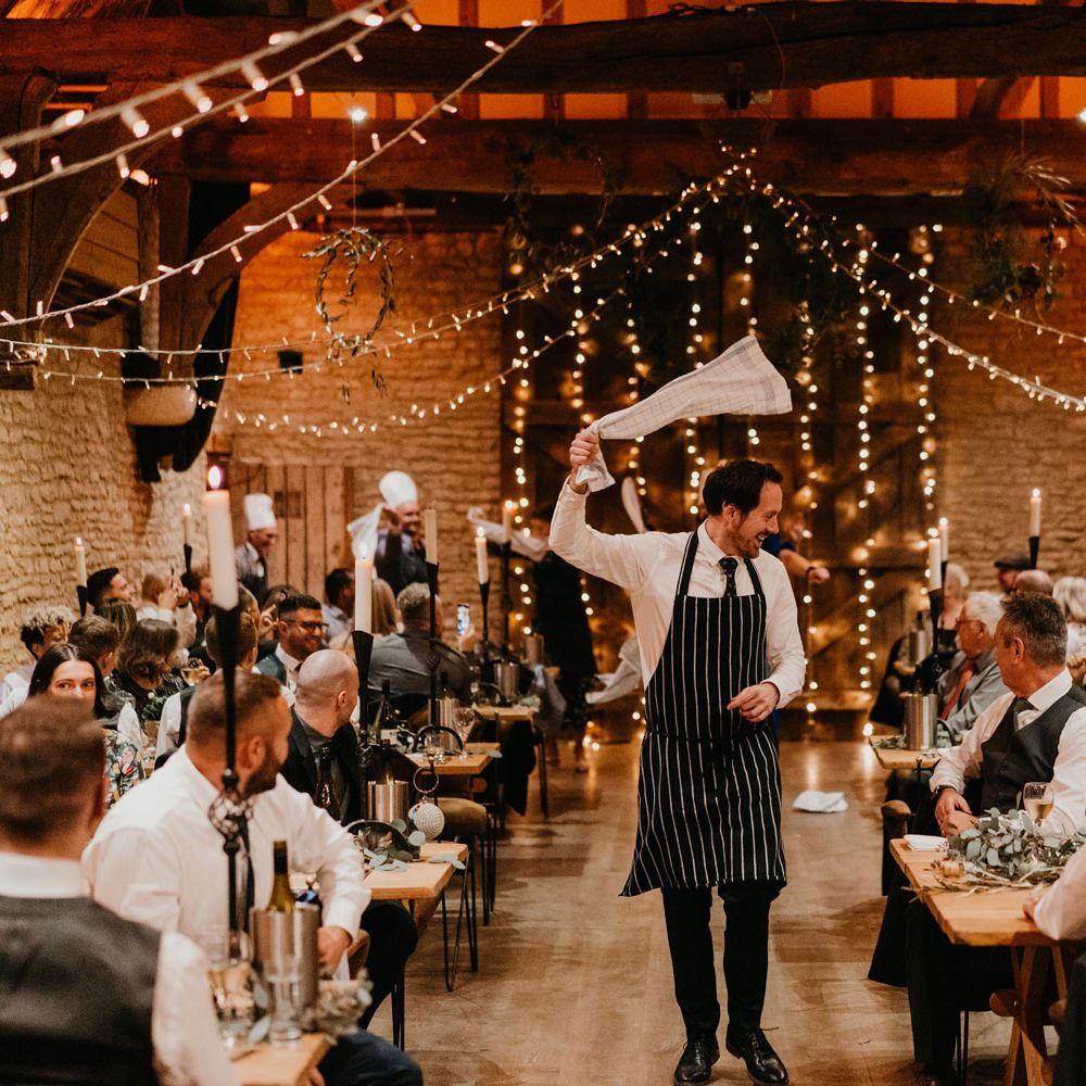 Server in striped apron swings white tea towel around as he walks down the middle of rustic wedding reception at The Tythe Barn 