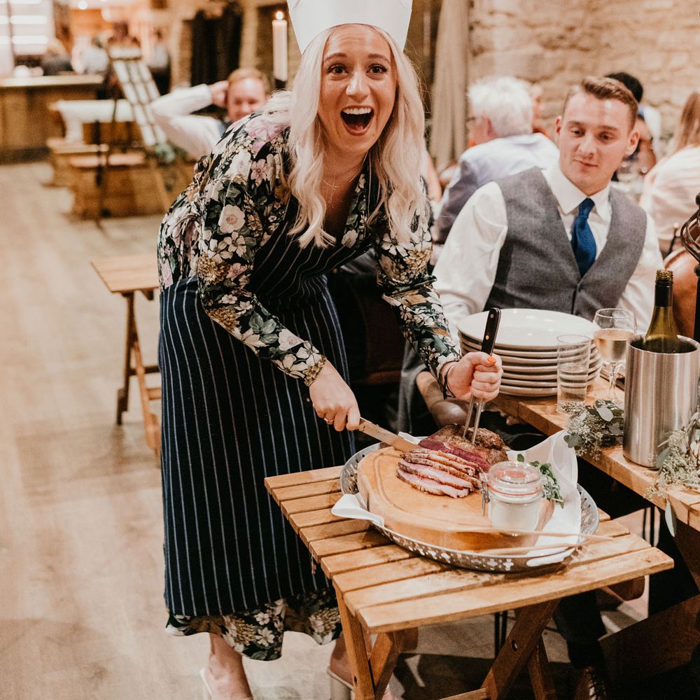 Guest in striped apron and chefs hat laughs as she cuts up meat for a guests table during rustic barn wedding breakfast