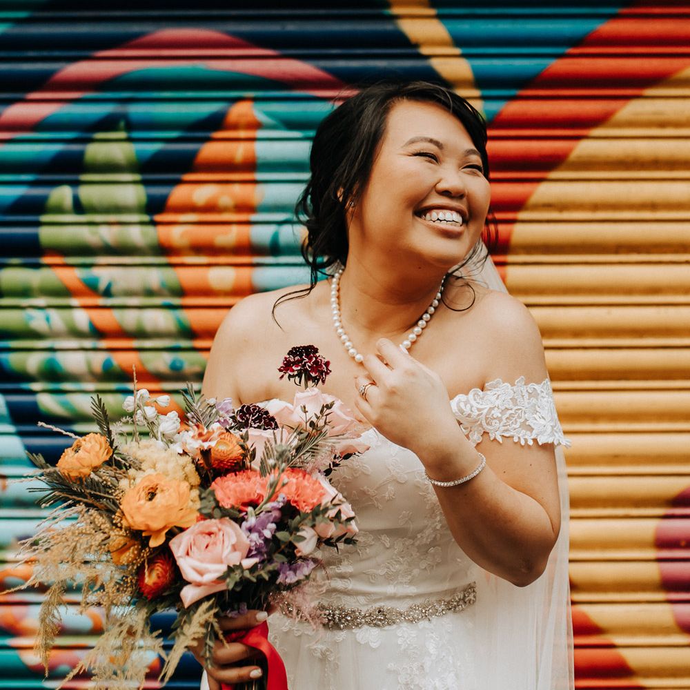 Laughing bride with pearl necklace and lace bardot wedding dress holding colourful wedding bouquet stands in front of street art by Scale wedding venue in Liverpool