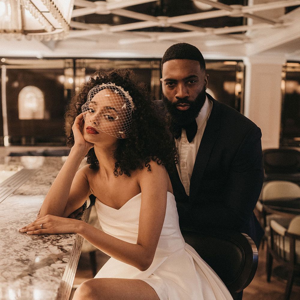 Black bride in a strapless wedding dress with front slit wearing a birdcage veil sitting at the bar with her groom in a tuxedo 
