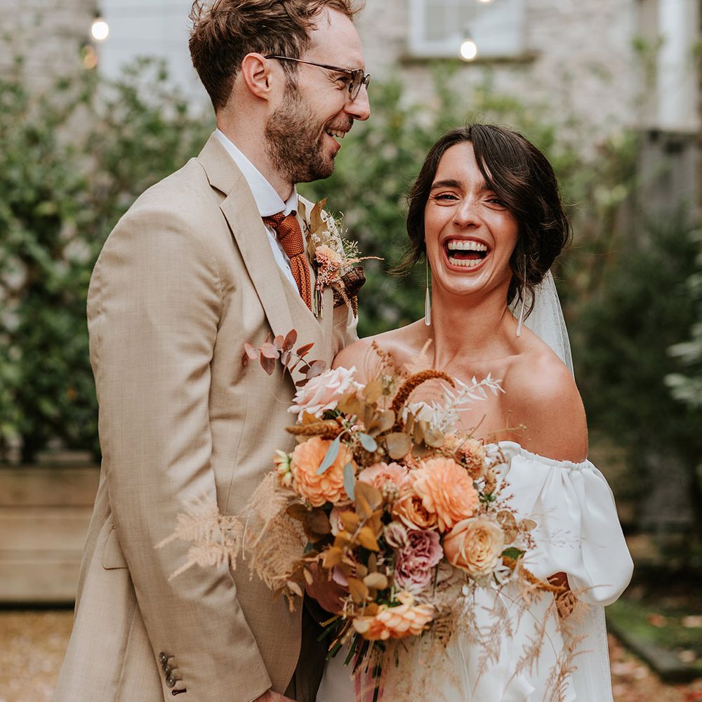 Laughing bride in puff sleeve wedding dress with groom in beige suit stand closely together