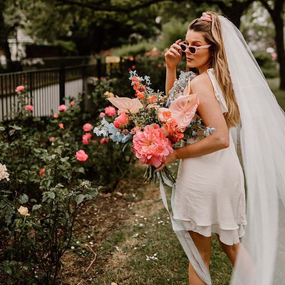 Stylish bride in a short wedding dress with retro sunglasses and long veil holding a pink and blue wedding bouquet 
