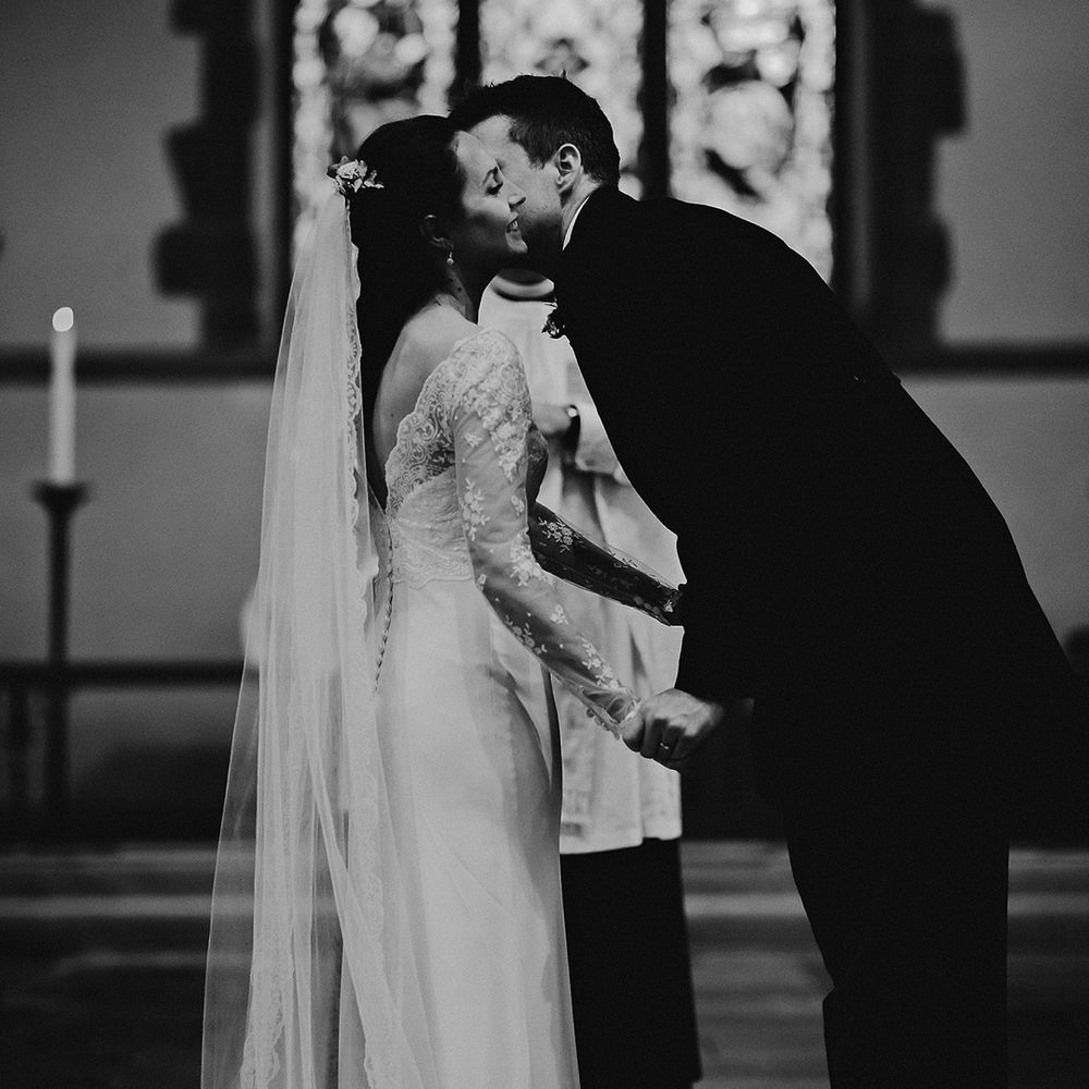 Groom leans in to kiss bride on the cheek with bride in white wedding dress with sheer lace sleeves and lace edge veil 