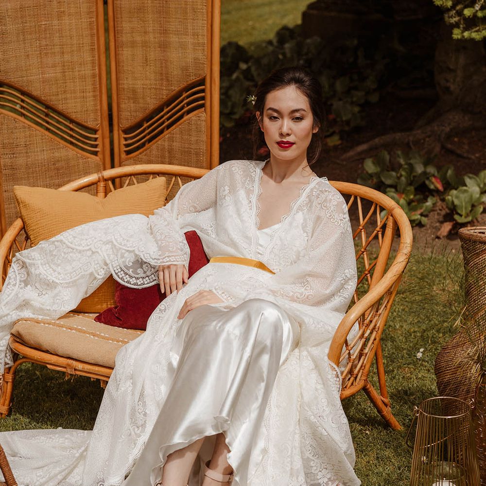 East Asian bride in a satin slip wedding dress with lace kimono style overlay sitting on a wicker chair at Japanese wedding shoot 