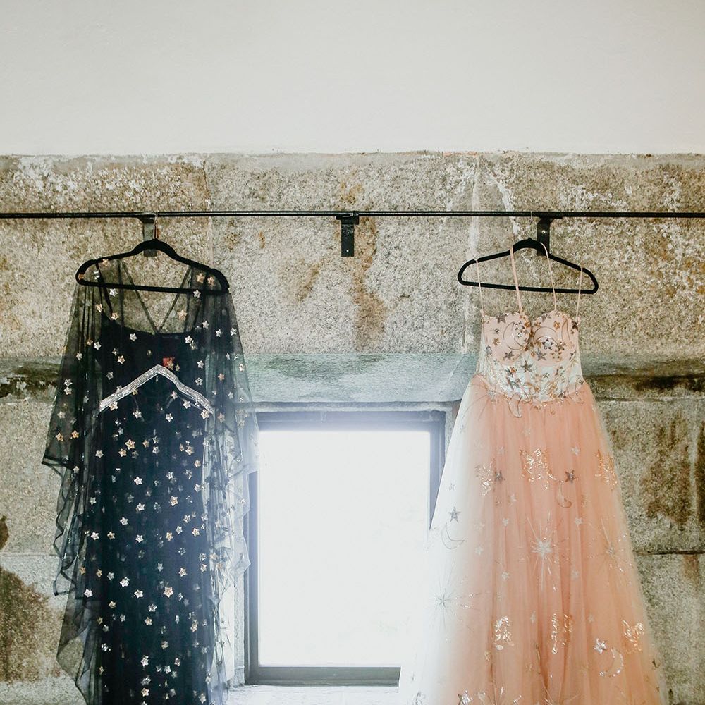 Bride's pink starry wedding dress and black celestial reception dress hang either side of window before wedding day