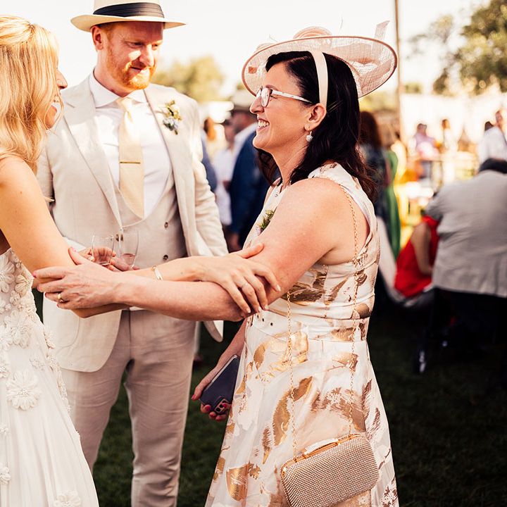 Bride in floral wedding dress and groom in cream suit and hat greets a wedding guest 