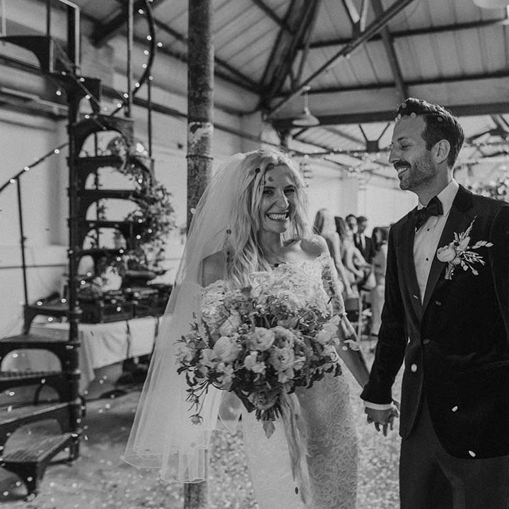 Bride in lace boho two piece with short veil and groom in black tux with bow tie and boutonniere standing amidst confetti at Hackney Studios
