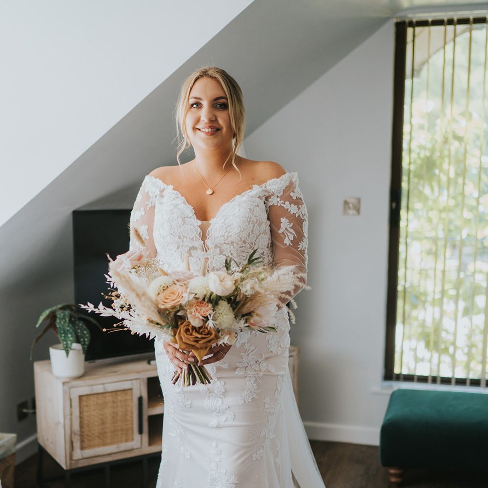 Bride wears lace long sleeve off the shoulder wedding dress and her blonde hair pulled into low up do 