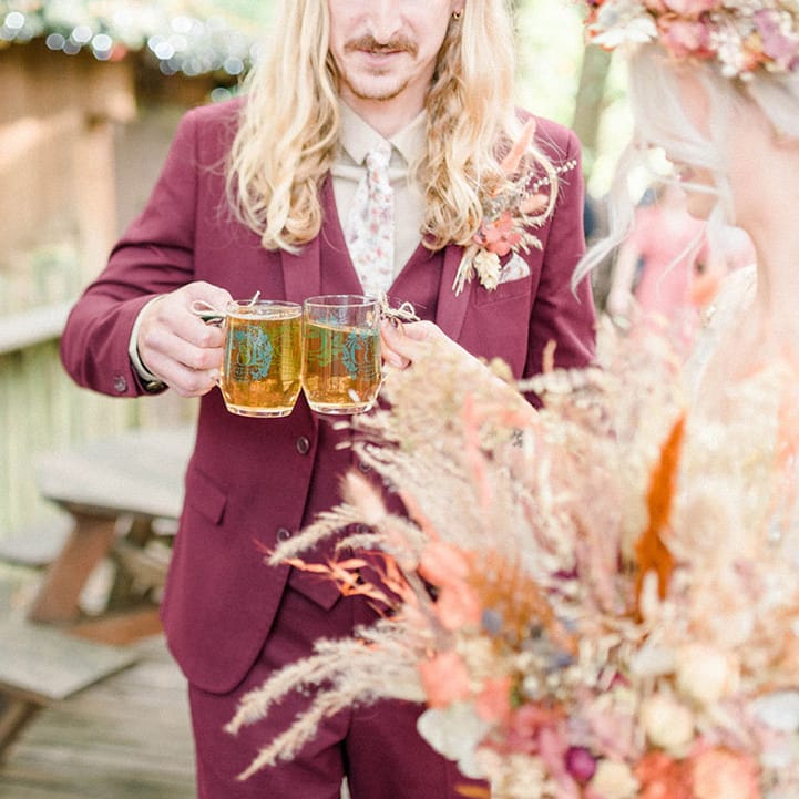 Groom in maroon suit and floral tie holds beer to his bride on their wedding day