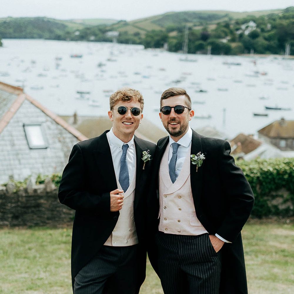Groom in sunglasses and his groomsmen wear morning suits with blue ties and floral buttonholes 