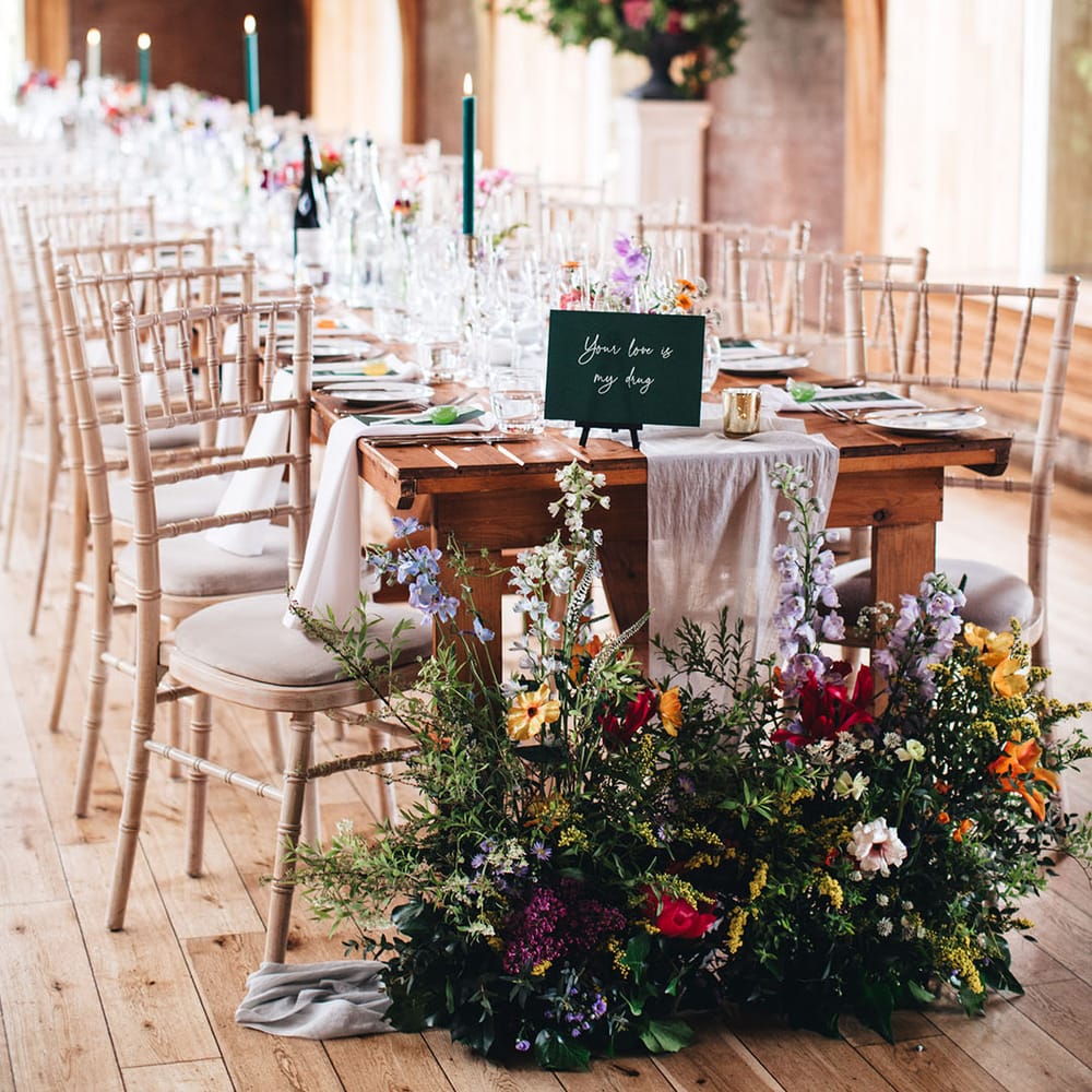 Lilac, yellow, red, and pink wedding wildflowers decorate the ends of the long banquet reception tables with green table name signs 