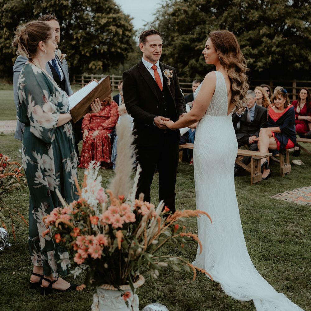 The bride and groom stand for their outdoor rustic glam ceremony with their guests sitting on benches at Rhyse Farm Worcestershire wedding venue 