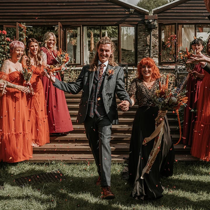 Groom in dark grey three piece suit and burgundy flower patterned tie and bride in embellished black wedding dress holding large orange dried flower bouquet 