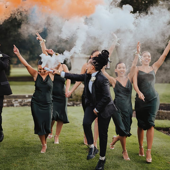 Bride in black snakeskin print tuxedo with black bowtie and white rose boutonniere running with wedding party in strappy satin midi length black bridesmaid dresses and classic black tuxedos with white rose boutonnieres doing smoke bomb wedding photos at Grittleton House wedding venue 