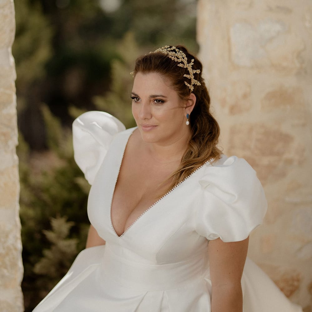 Bride in white satin dress featuring puff sleeves and a plunge neckline with pearl earrings and a gold leaf bridal crown