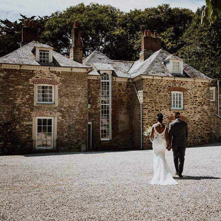 Bride in v back lace and mesh wedding dress holding hands with groom in charcoal grey grooms suit walking around the grooms of Tredudwell Manor wedding venue