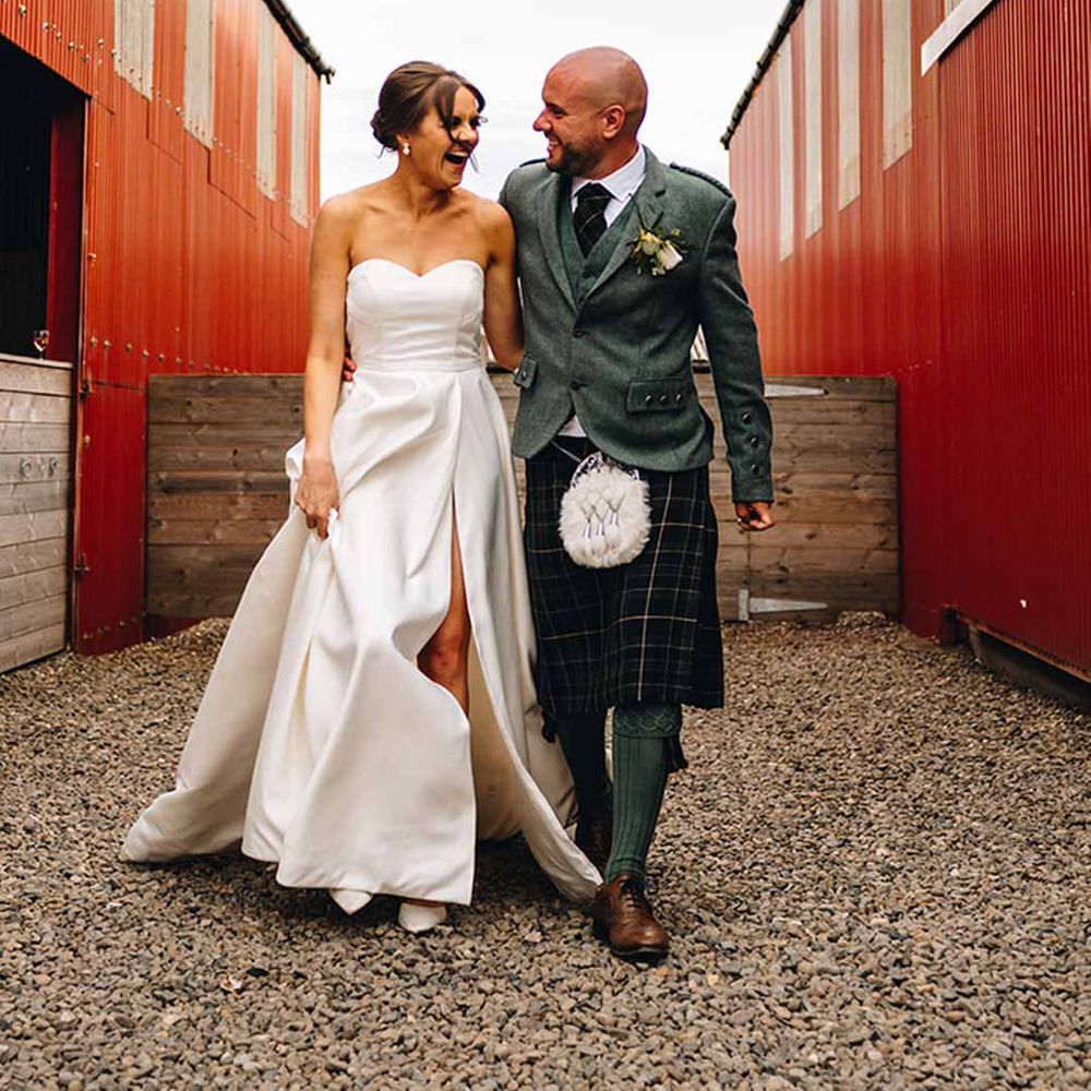 Bride in strapless satin wedding dress with front slit walking with groom in wedding kilt and sporran at Dalduff Luxury Barn Weddings Scotland