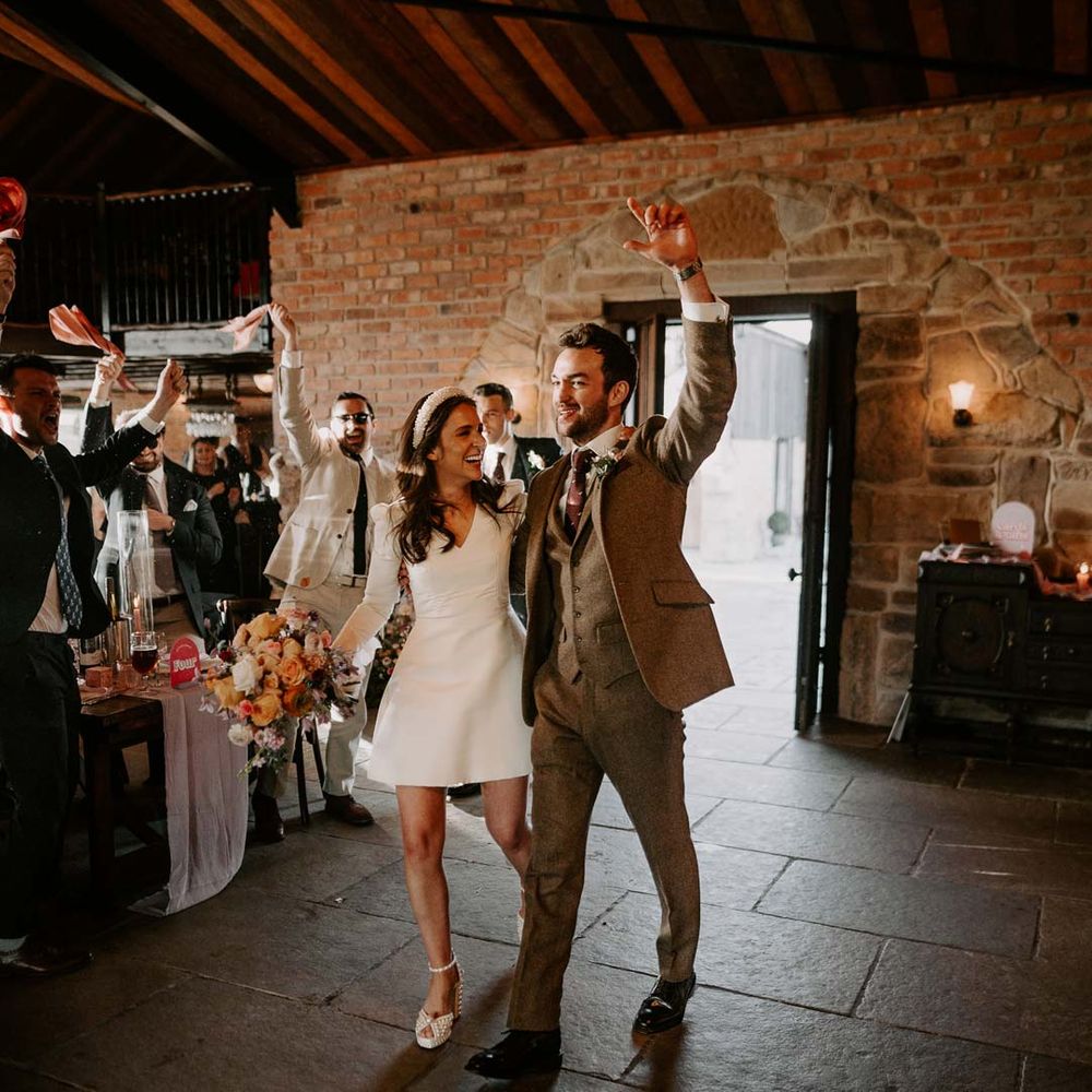 Bride in long sleeve short wedding dress, pearl bridal headband and platform pearl wedding heels walking into reception room of Willow Marsh Farm Loughborough with groom in three piece chocolate brown grooms suit 
