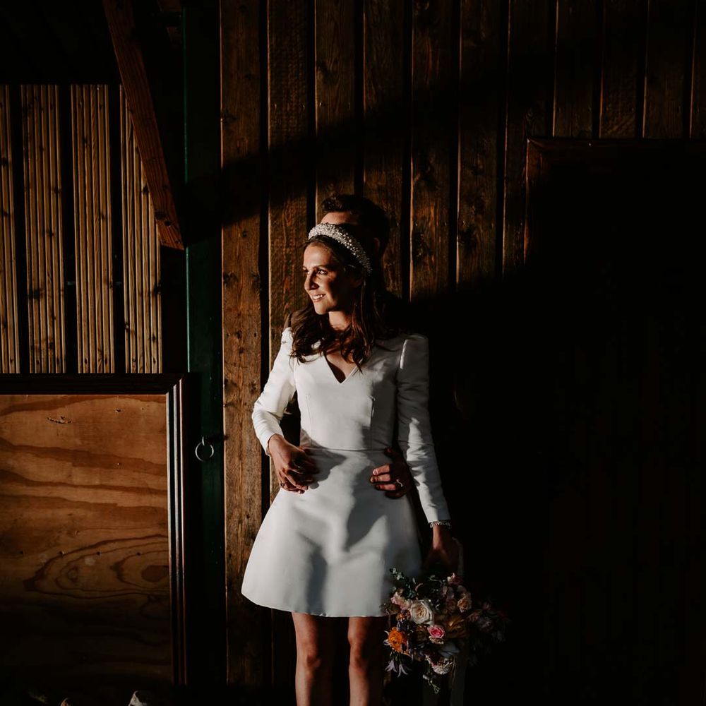 Bride standing in the shadows in long sleeve short wedding dress, pearl bridal headband and platform pearl wedding heels with groom behind her