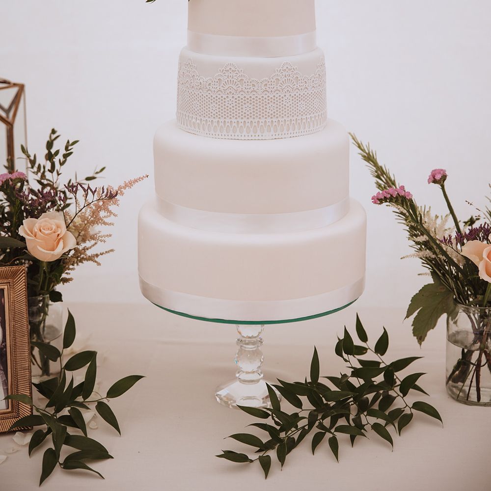 Four tier white iced traditional wedding cake with white ribbon and lace with pink flower decorations 