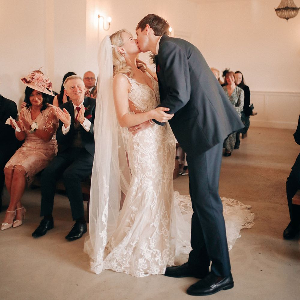 Bride in mermaid style fitted lace wedding dress with embroidered veil kissing groom at ceremony 