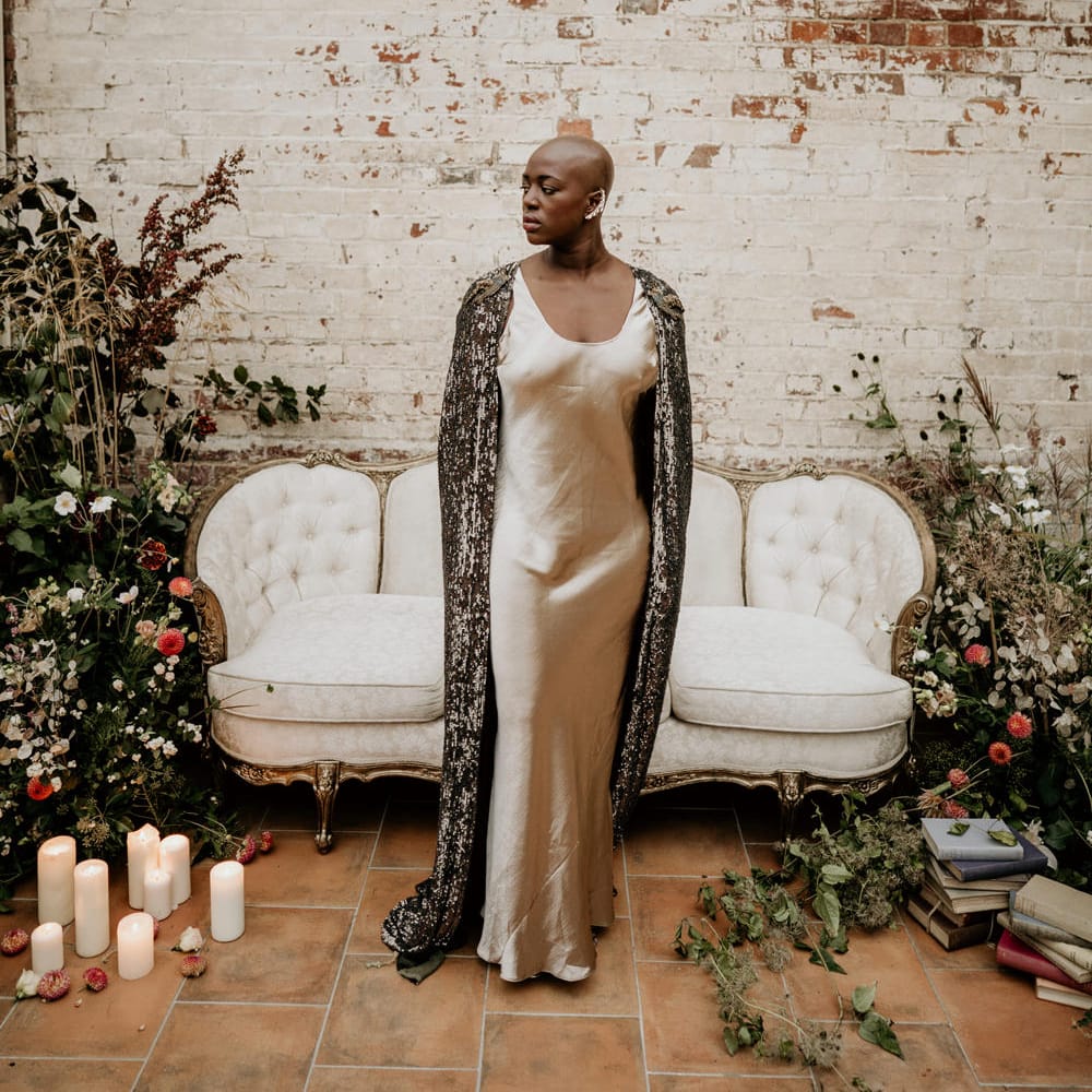 Black bride with alopecia in a beige satin dress and black shimmer cape wearing a gold ear cuff 