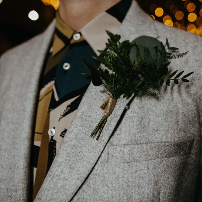 Groom in a grey suit and patterned shirt wearing a green foliage buttonhole
