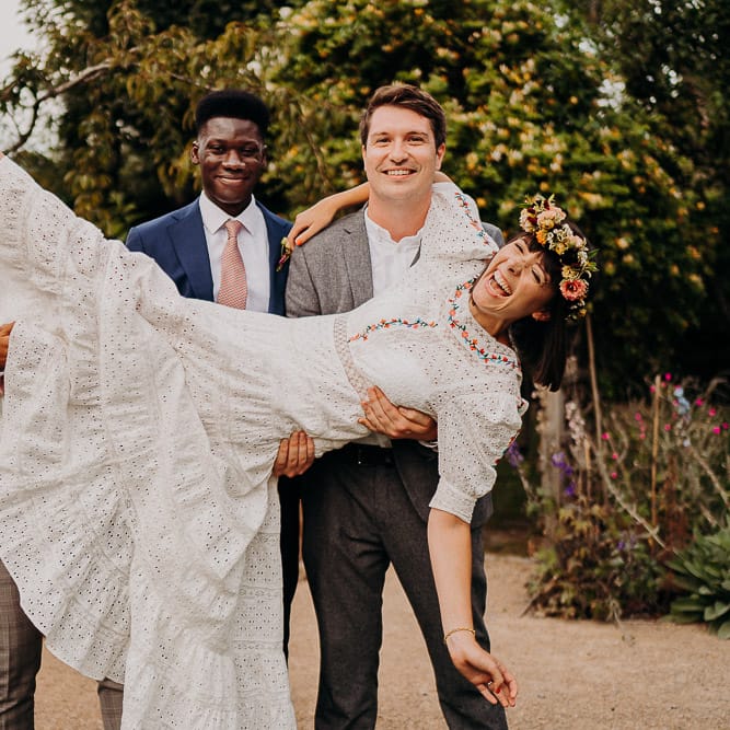 Groom and groomsmen picking up the bride in a white dress with colourful embroidery, flower crown and red shoes 
