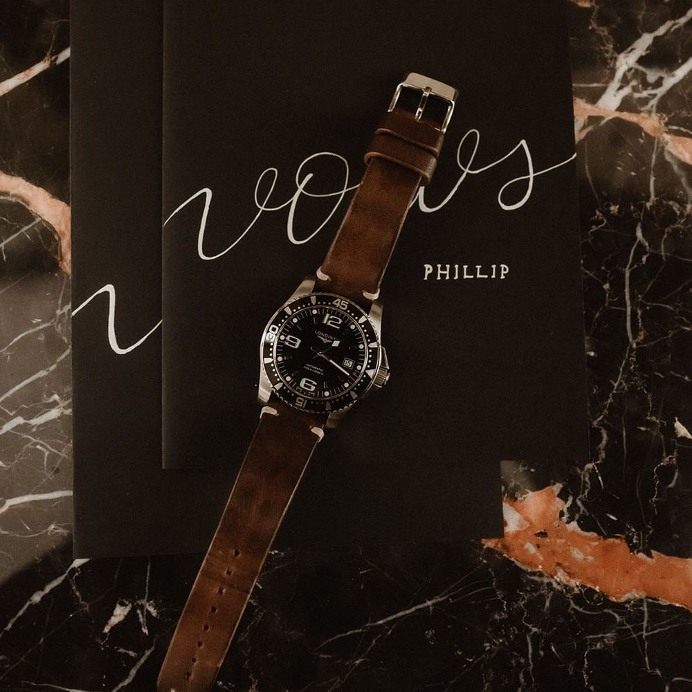 Men's watch with a brown strap, laid on top of black wedding vows booklets with calligraphy font