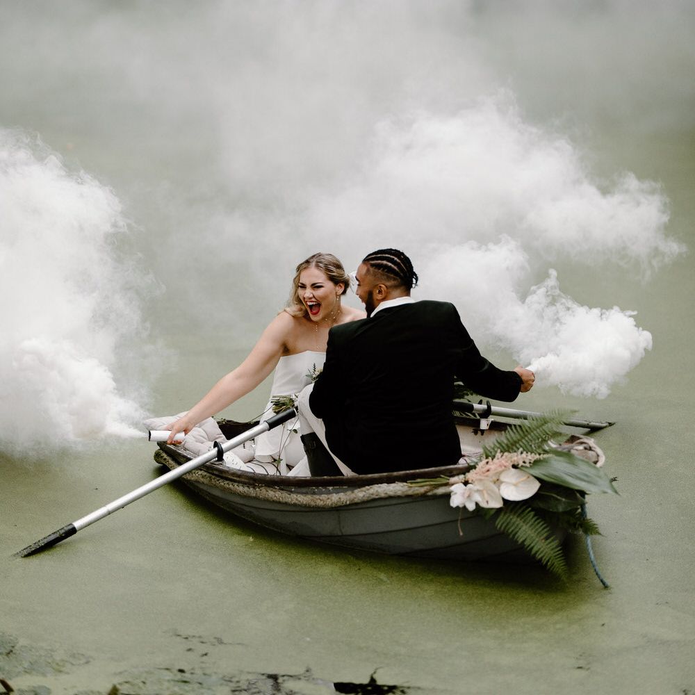 Bride and groom on a green algae covered lake in a row bow, holding white smoke flares
