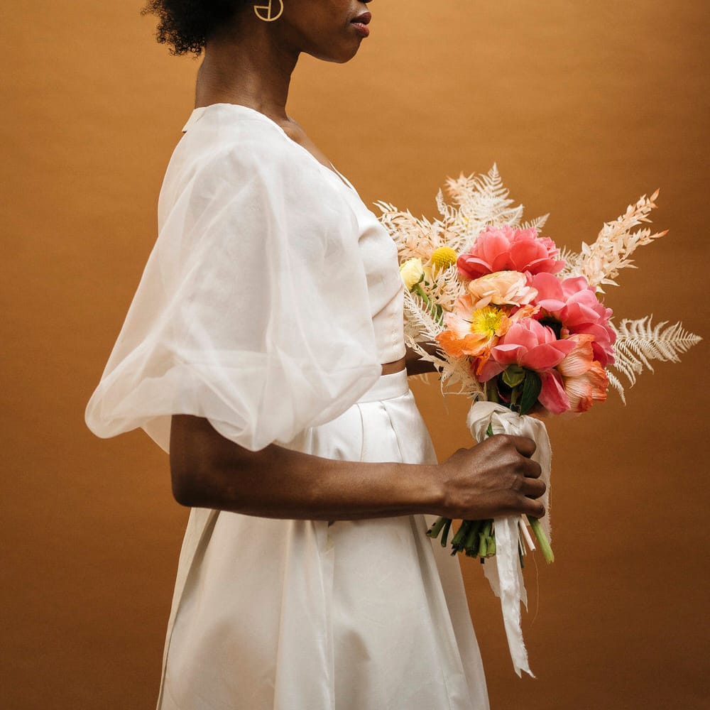Bride wearing fresh flowers in her afro, carrying a matching bouquet and wearing a puff sleeve wedding top