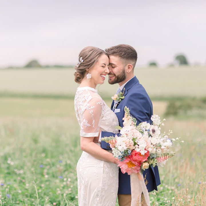 Bride and groom laughing in a field with the bride in a lace wedding dress holding a wildflower wedding bouquet and the groom in a blue blazer 