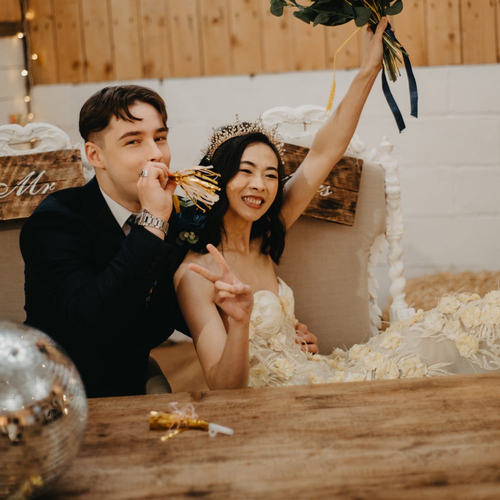 Bride in strapless feathered wedding dress holds up peace sign and tropical wedding bouquet as groom blows party blower with streamers at rustic wedding reception