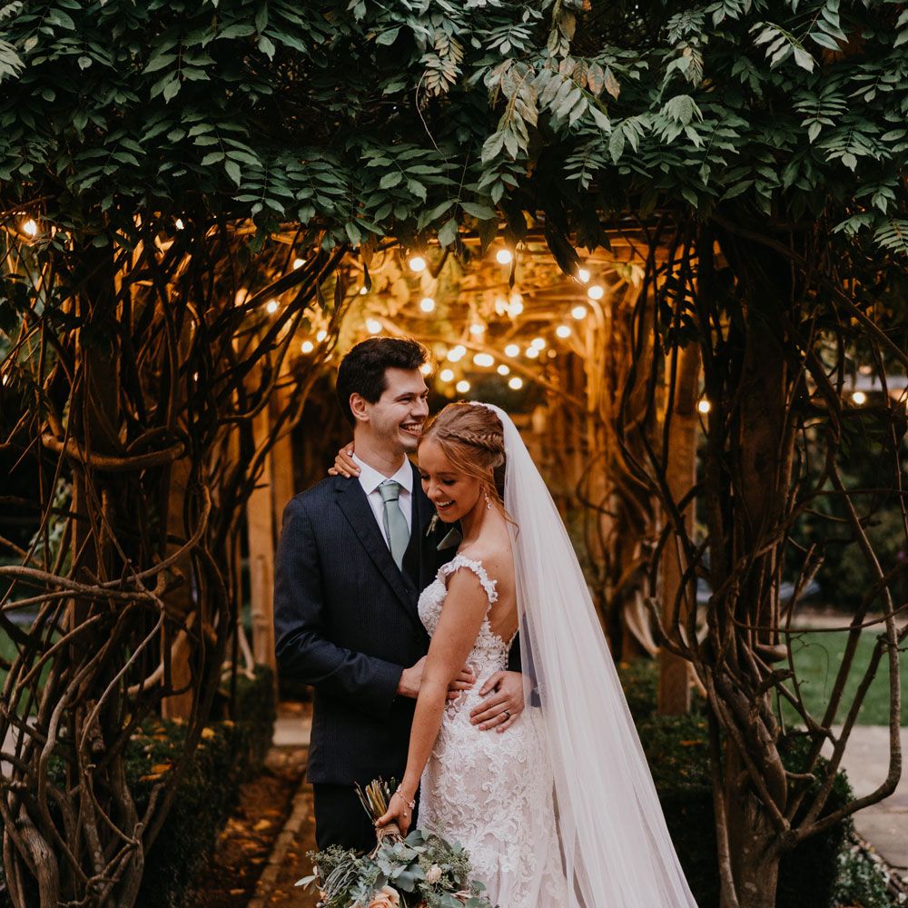 Bride in open back lace and tulle wedding dress and veil stands hugging groom in dark suit in the grounds of The Tythe Barn in Launton