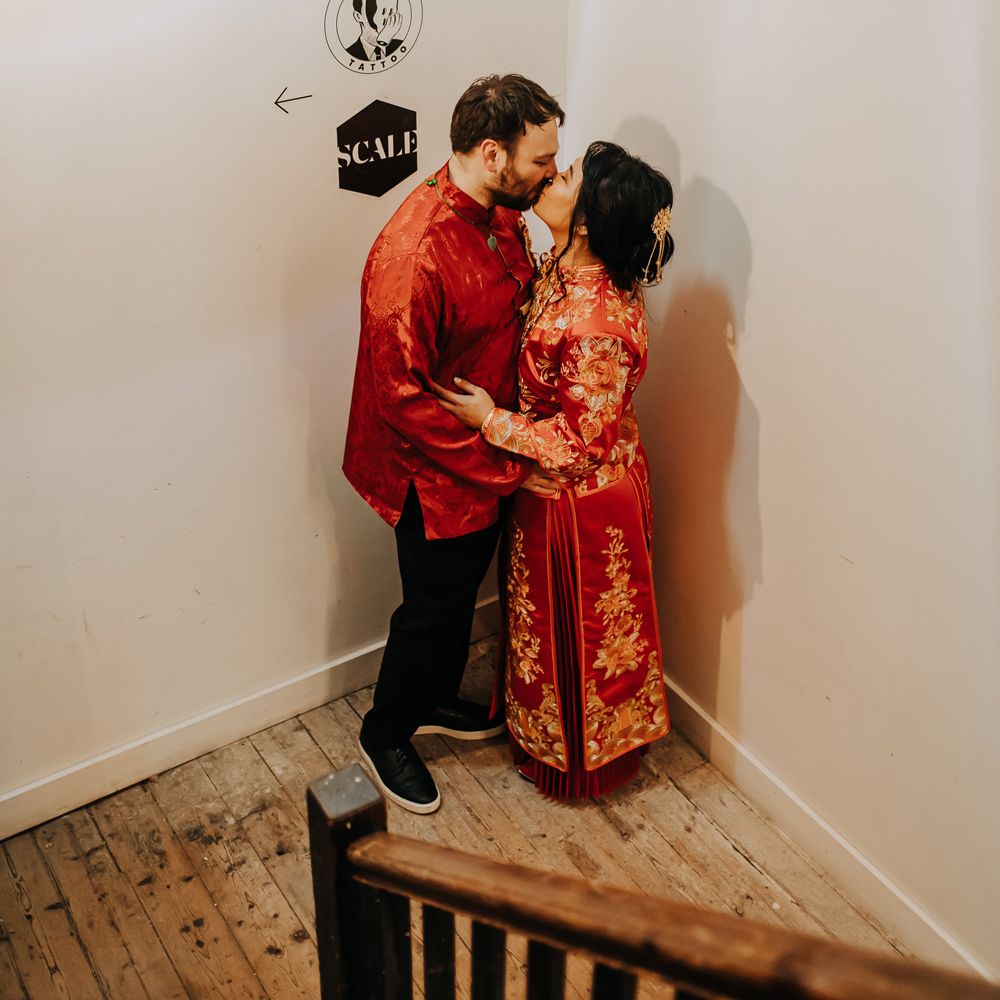 Bride in red and gold traditional Chinese tea ceremony dress kisses groom in black trousers and red traditional Chinese clothing