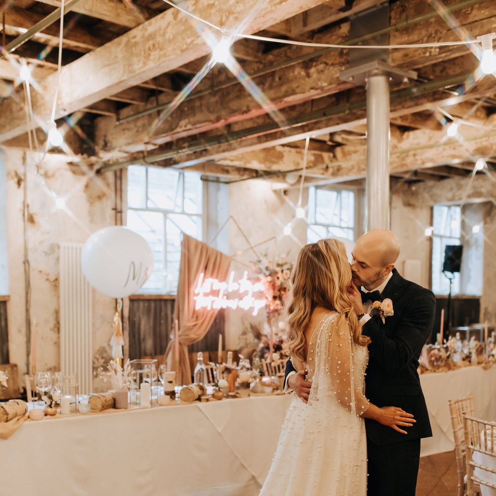 Bride in white wedding dress with train and pearl details kisses groom in navy suit as they stand in Holmes Mill wedding reception room with neon wedding sign