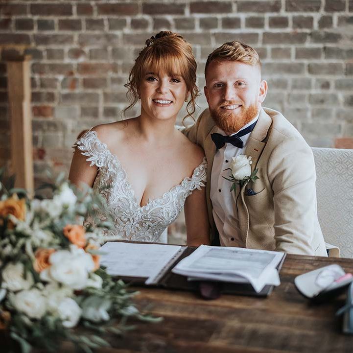 Bride and groom smile together as they sign the register with white and orange colour scheme for spring wedding