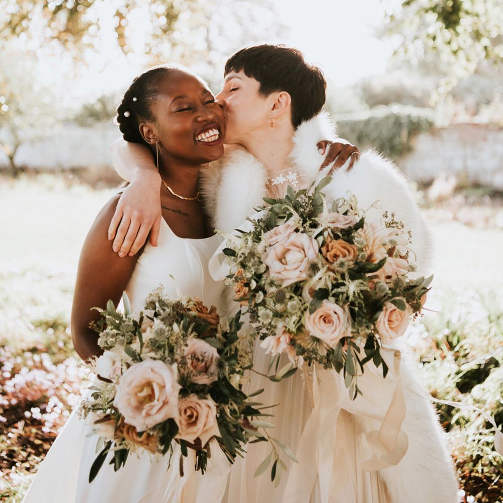 Bride with short hair kissing her bride under a tree at Elmore Court holding romantic pink wedding bouquets 
