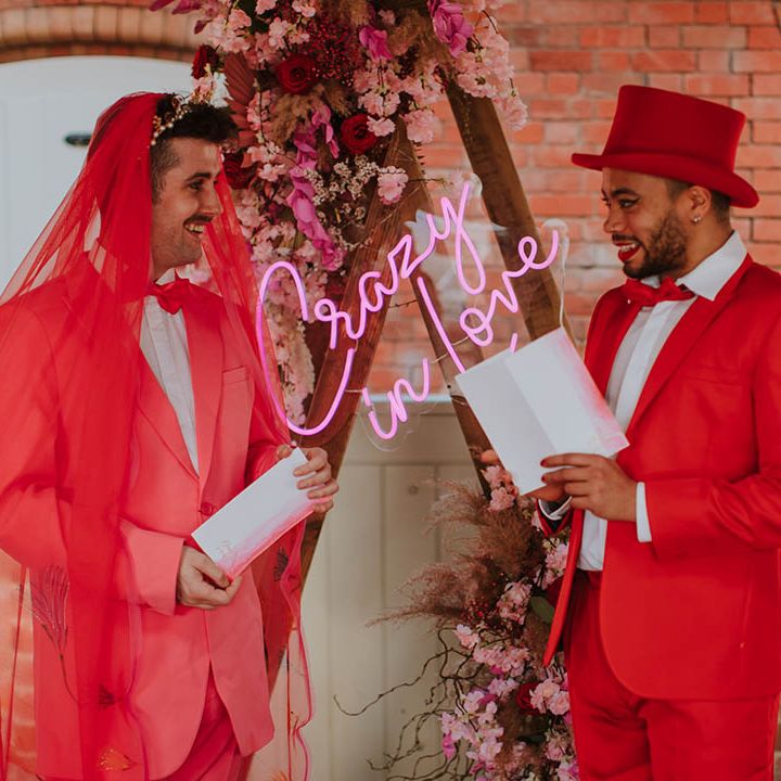 LGBTQI+ couple exchanging vows at Prestwold Hall Barns in a pink suit with red veil and a red suit and top hat 