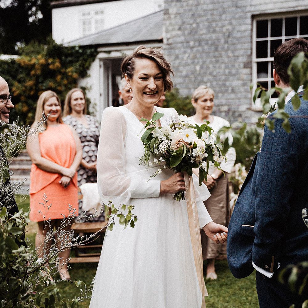 Bride smiles at the altar for outdoor wedding ceremony at Treseren holding white flower bouquet including cosmos and dahlias