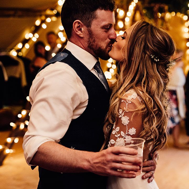 Bride and groom kiss in front of fairy light as the groom holds a beer 