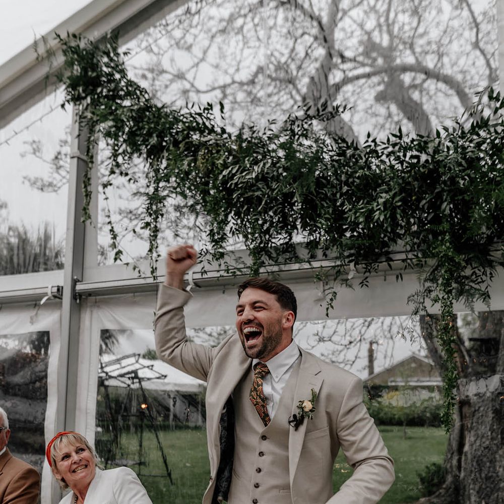 Groom in a stone coloured three piece with paisley tie fist bumping he air during the clear marquee wedding reception  