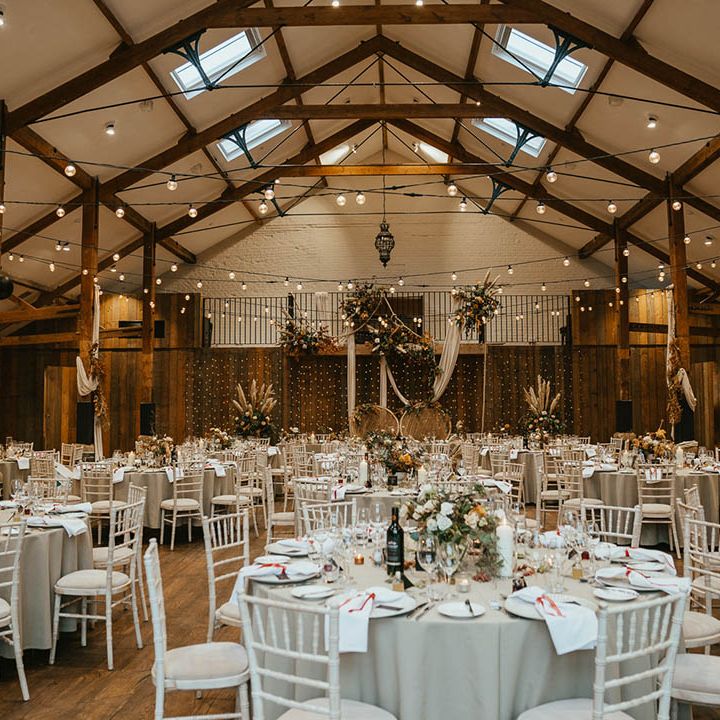 Grey wedding table settings with venue decorated with festoon lighting, pampas grass, peacock chairs and white wedding drapes 