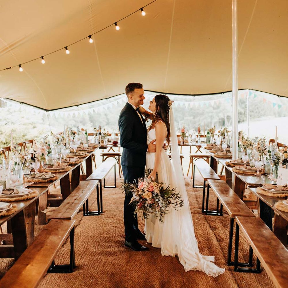 Bride in sleeveless wedding dress with long veil and large neutral bouquet and groom in dark suit standing in DIY tent at garden wedding