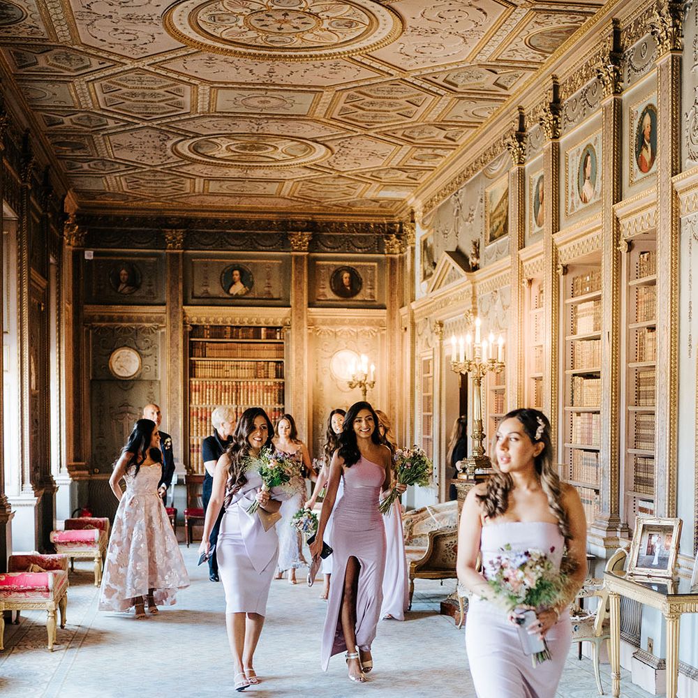 Bridesmaids wearing lavender bridesmaid dresses and holding brightly coloured floral bouquet walk through grand room at Syon Park