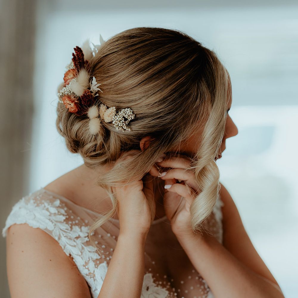 Bride has her blonde hair in an updo with bunny grass hair accessories as she puts in earrings 