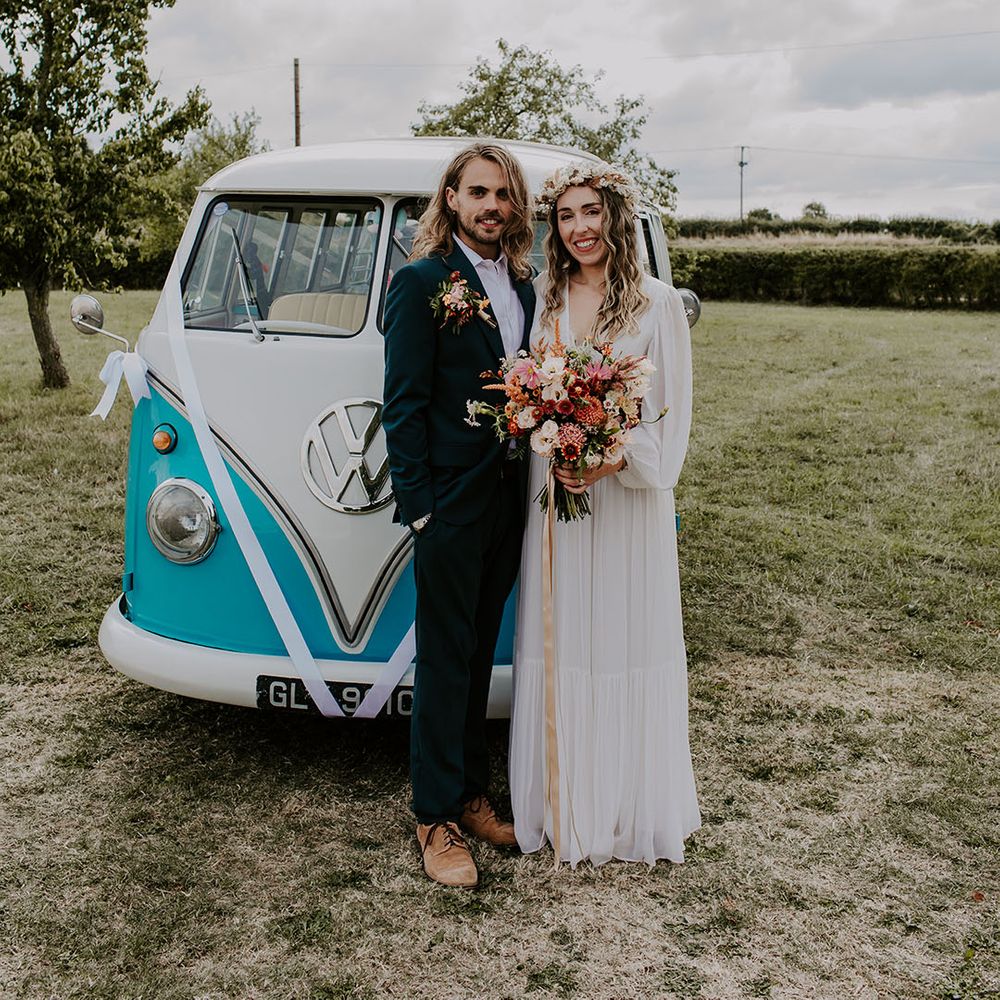 Bride & groom stand in front of VW camper van as bride holds colourful floral bouquet tied with champagne coloured ribbon