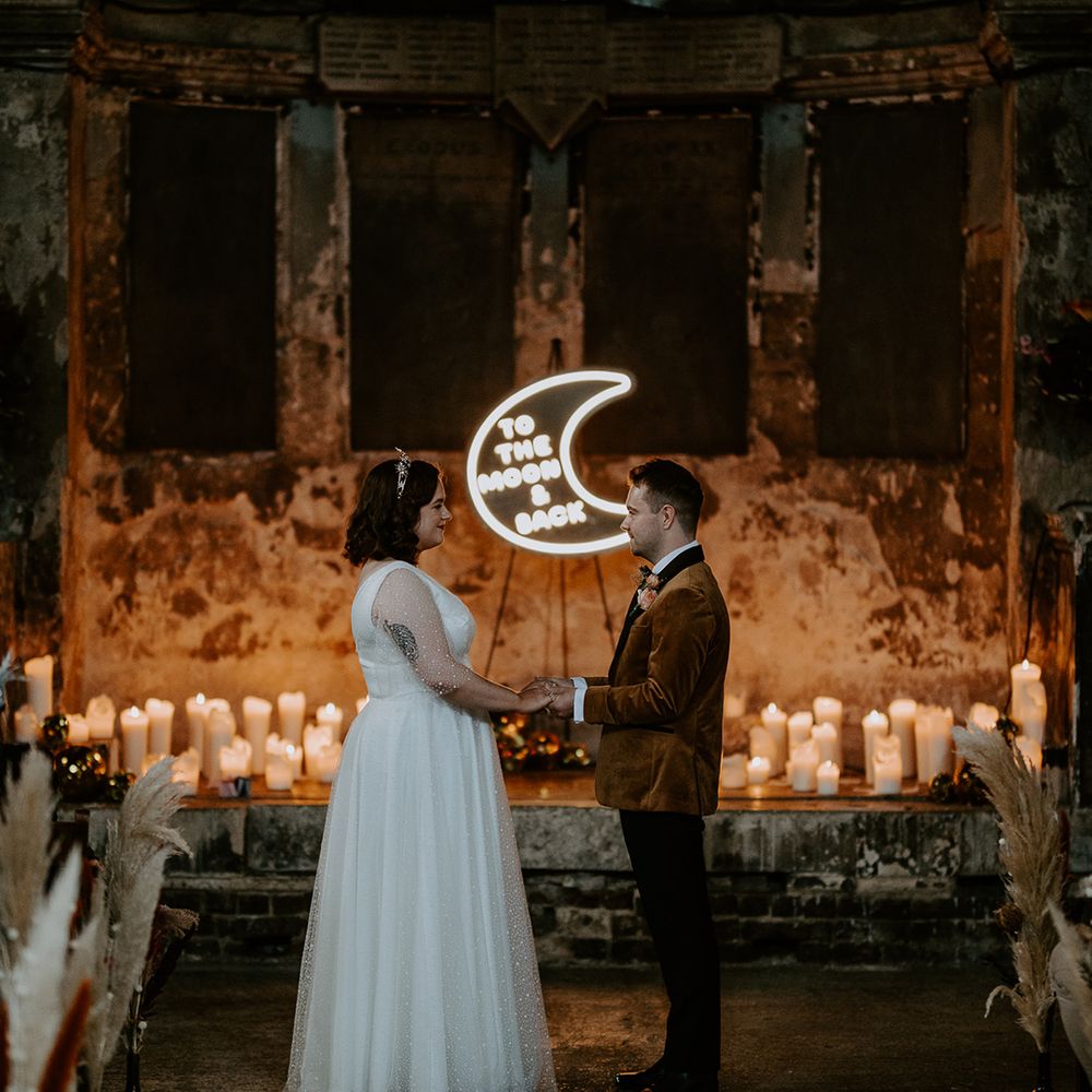 The couple look lovingly at each other surrounded by warming wedding lighting created by candles and a moon neon sign