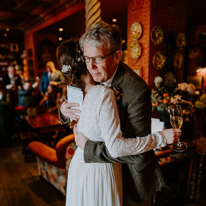 Bride holding a Champagne glass wearing long sleeve lace vintage wedding dress with drop back detail, pearl earrings and pearl hair accessories hugging her dad at intimate London wedding