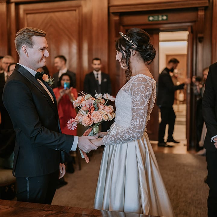 Groom in classic black tux with bowtie and rose boutonniere holding hands with bride in  taffeta material princess wedding dress with long lace sleeves and gold, pearl and emerald bridal crown holding bouquet with light pink poppies, orange garden roses, eucalyptus, Christmas red berry garlands, dried flowers and sage green and orange ribbon